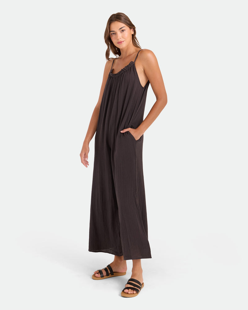 A woman poses against a white background wearing the Volcom Women's Ruffle Up Romper, a loose black sleeveless jumpsuit with thin straps and wide legs. She has one hand in her pocket and pairs it with black and tan flat sandals.