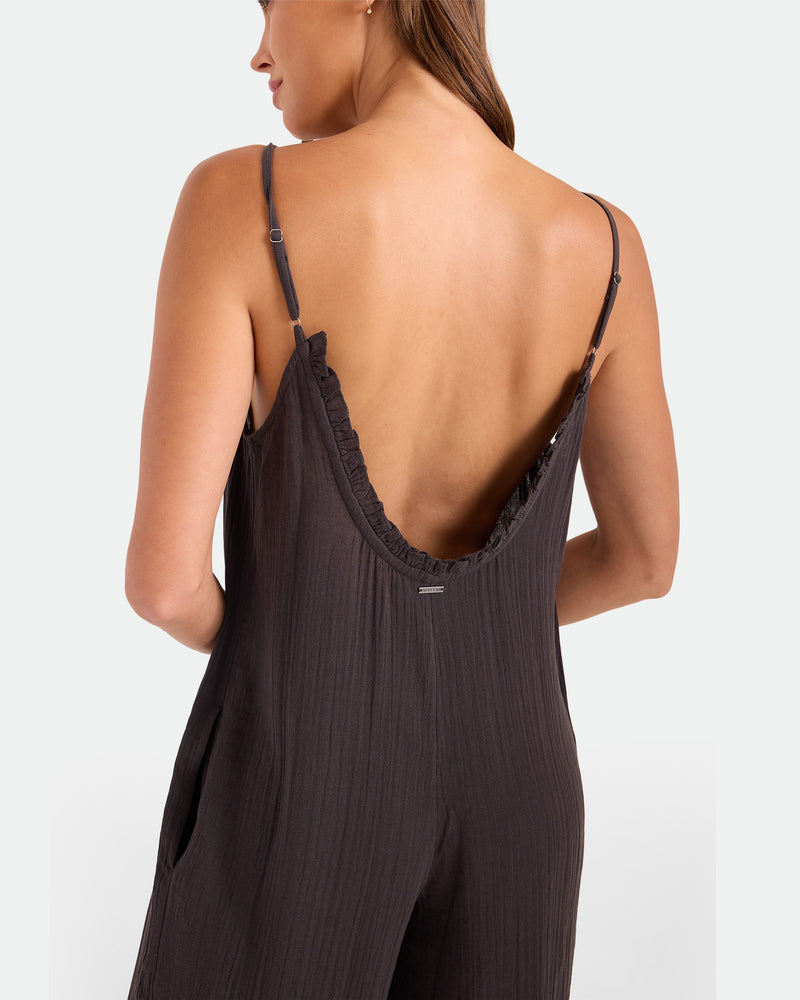 A woman is seen from behind wearing the Volcom Women's Ruffle Up Romper, a loose, dark jumpsuit with thin straps and a deep back. The textured fabric complements her long hair, which drapes over one shoulder.