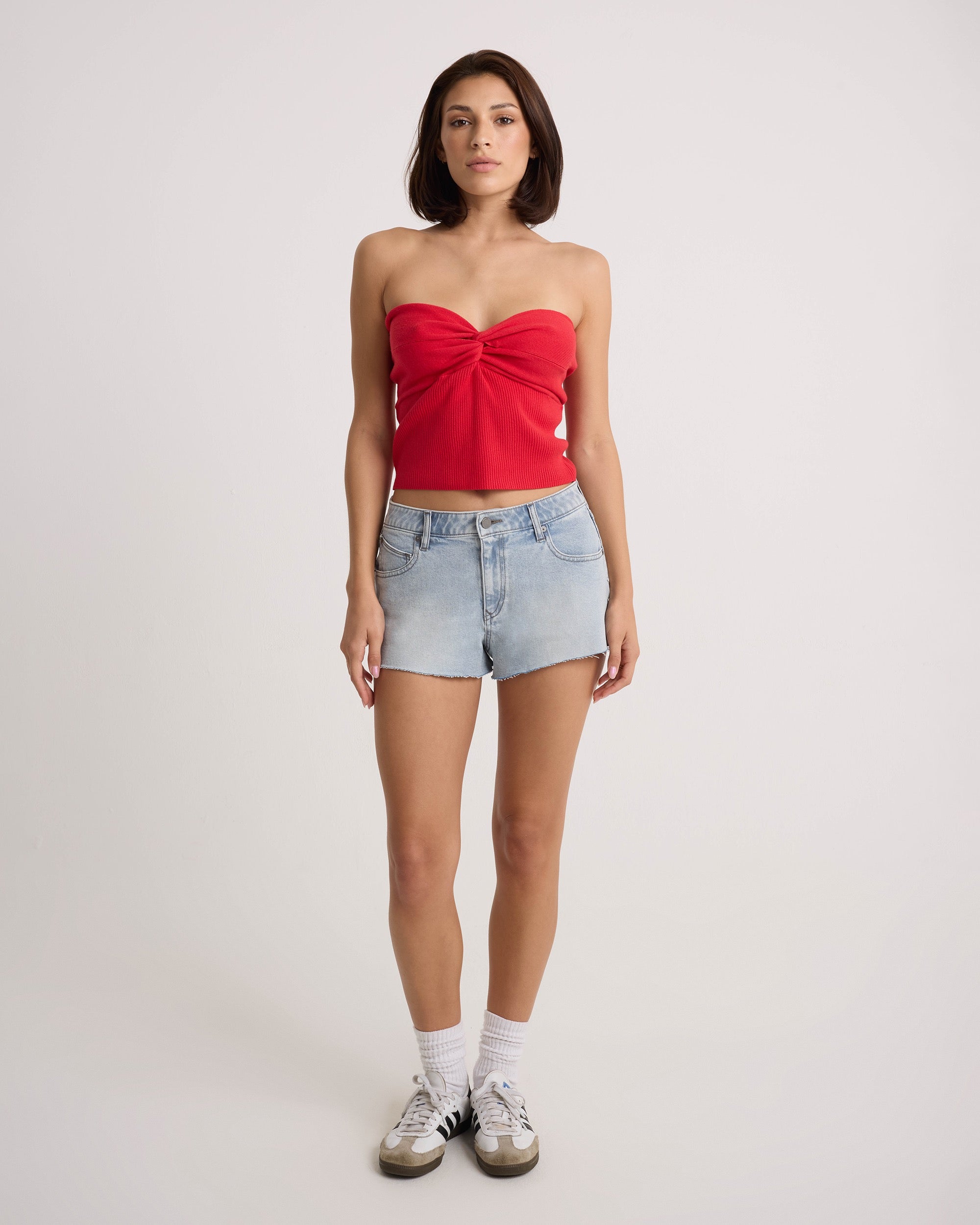 A person stands facing forward in the Volcom Women's Cruz Control red strapless top and light blue high-rise chambray-wash denim shorts, with white socks and black-and-white sneakers, against a plain white background.