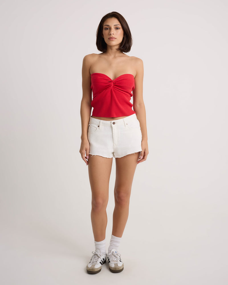 A woman faces forward wearing the Volcom Women's Cruz Control red strapless top and vintage white high-rise denim shorts, styled with white socks and sneakers, against a plain light background.
