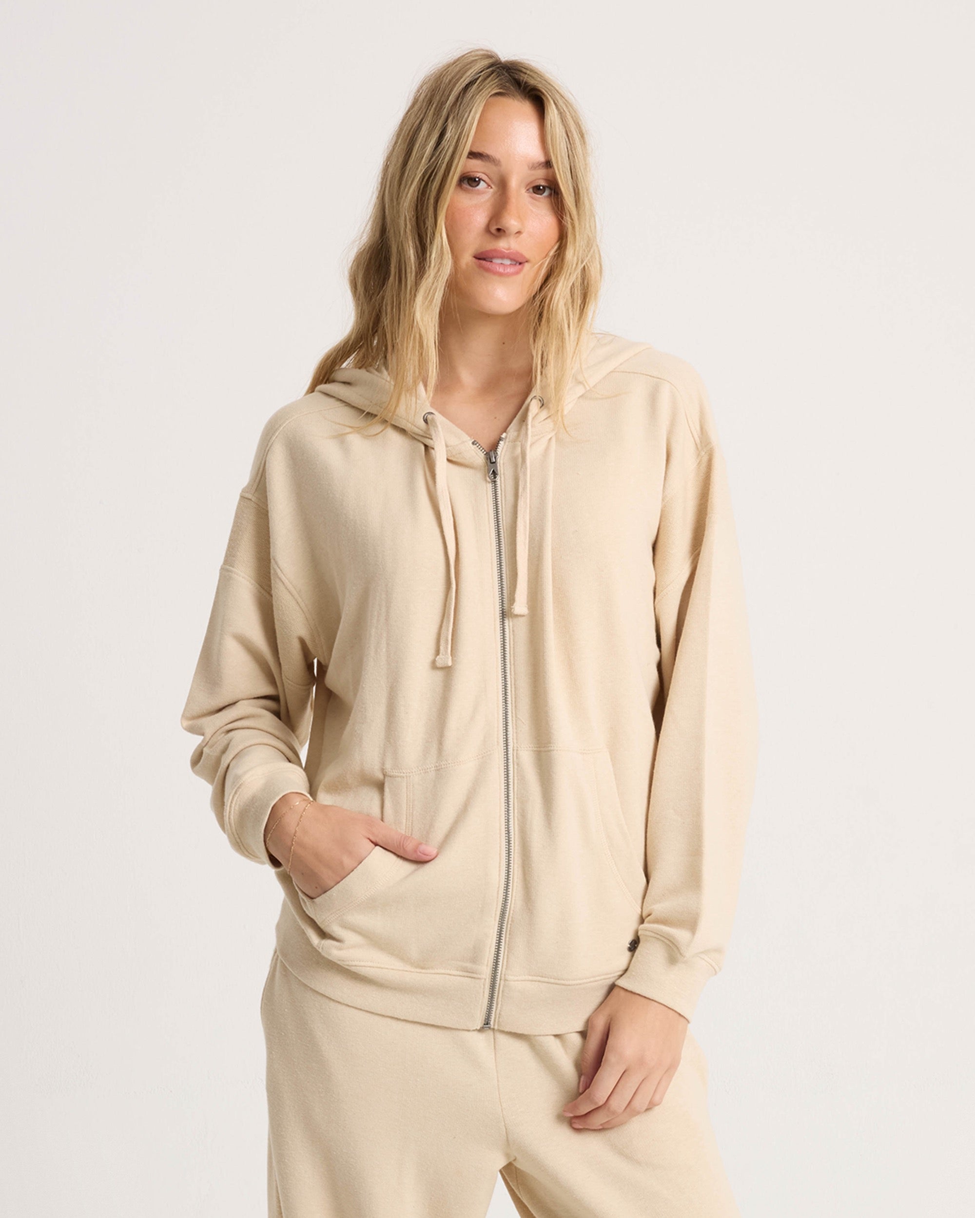 A woman with long blonde hair wears the Volcom Women's Lil Frenchie Zip Hoodie, standing against a plain white background, facing forward with one hand in her pocket.
