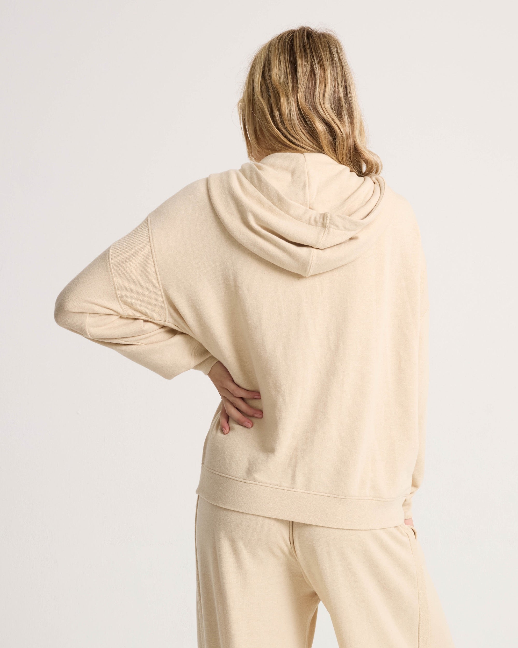A person with long, blonde hair stands with their back to the camera, wearing a light beige Volcom Women's Lil Frenchie Zip Hoodie and matching pants. Their right hand rests on their hip against a plain white background.
