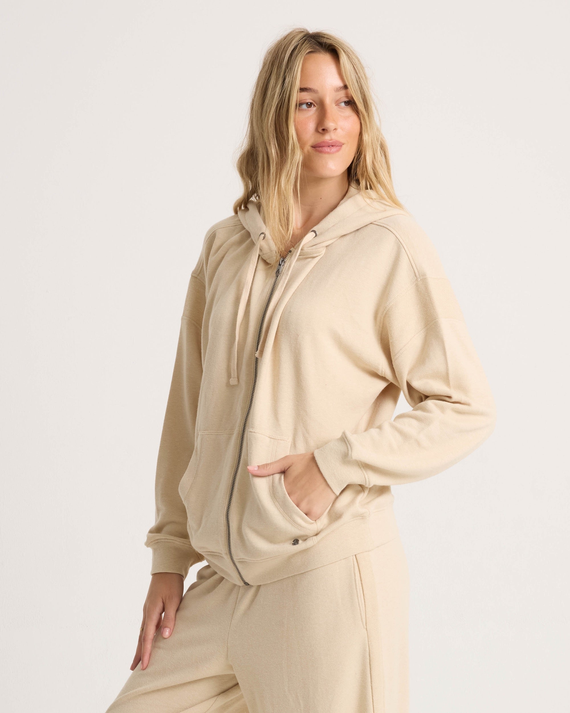 A woman with long blonde hair wears the Volcom Women's Lil Frenchie Zip Hoodie in beige and matching sweatpants, standing against a plain light background with one hand in her pocket, looking slightly to the side.