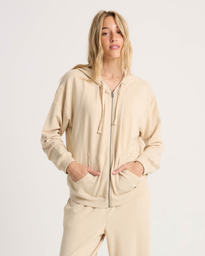 A person with long, blonde hair wears the Volcom Women's Lil Frenchie Zip Hoodie in beige, with matching pants, standing against a plain white background and hands in the front pockets of the cozy sweatshirt.