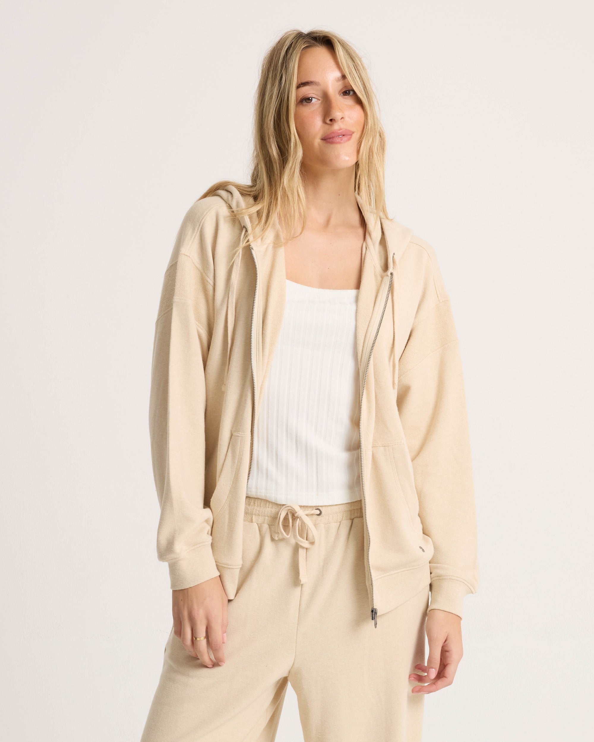 A woman with long blonde hair wears the Volcom Women's Lil Frenchie Zip Hoodie in beige and matching drawstring pants over a white ribbed tank top, standing against a plain light background and smiling at the camera.