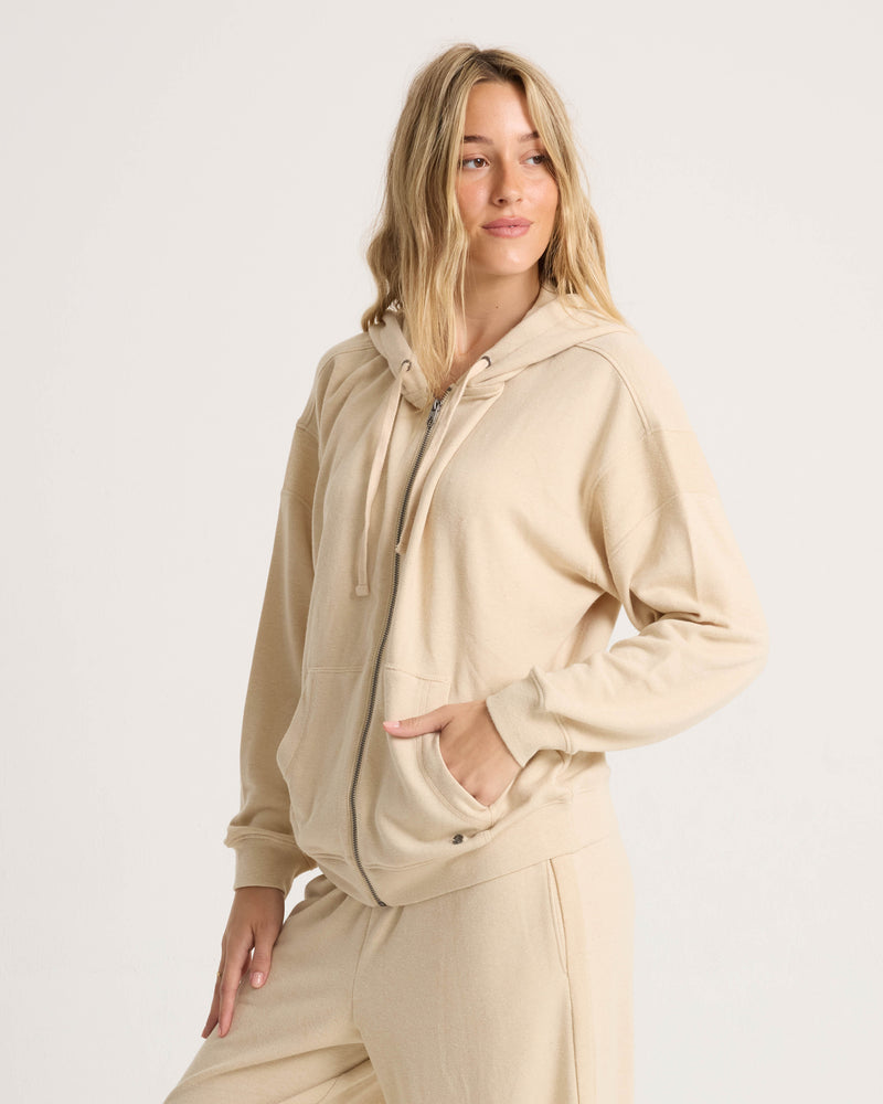 A woman wearing the Volcom Women’s Lil Frenchie Zip Hoodie in a relaxed beige fit, paired with matching sweatpants, stands with one hand in her pocket against a plain light background.