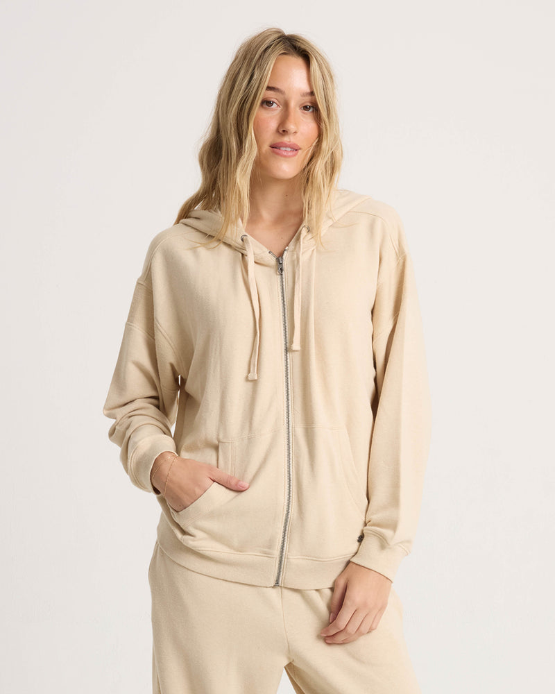 A woman with long blond hair stands against a plain background, wearing the Volcom Women’s Lil Frenchie Zip Hoodie and matching pants. The relaxed-fit fleece hoodie is fully zipped, and she has one hand in her pocket with a laid-back expression.