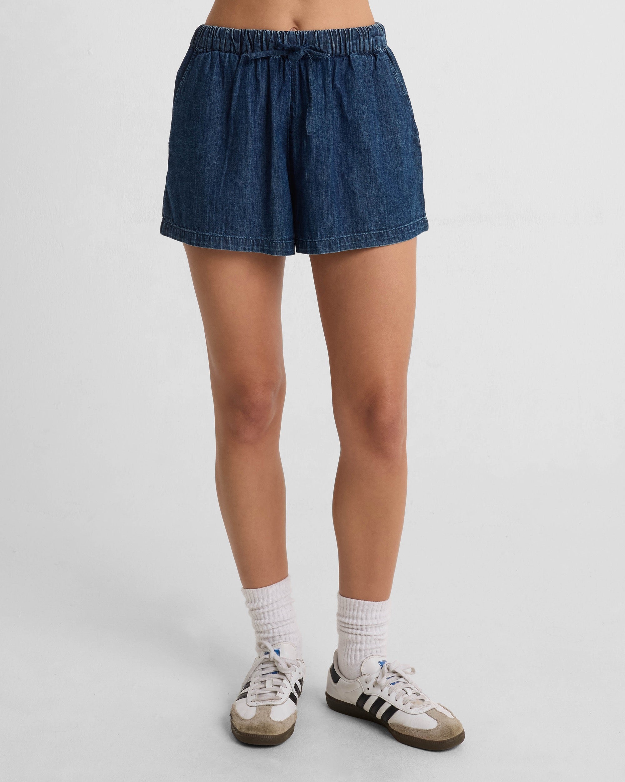 A person wears Volcom Women's Stone Shine Shorts, made from 100% cotton, with white socks and white sneakers featuring black stripes, standing against a plain white background. Only the lower half of the body is visible.