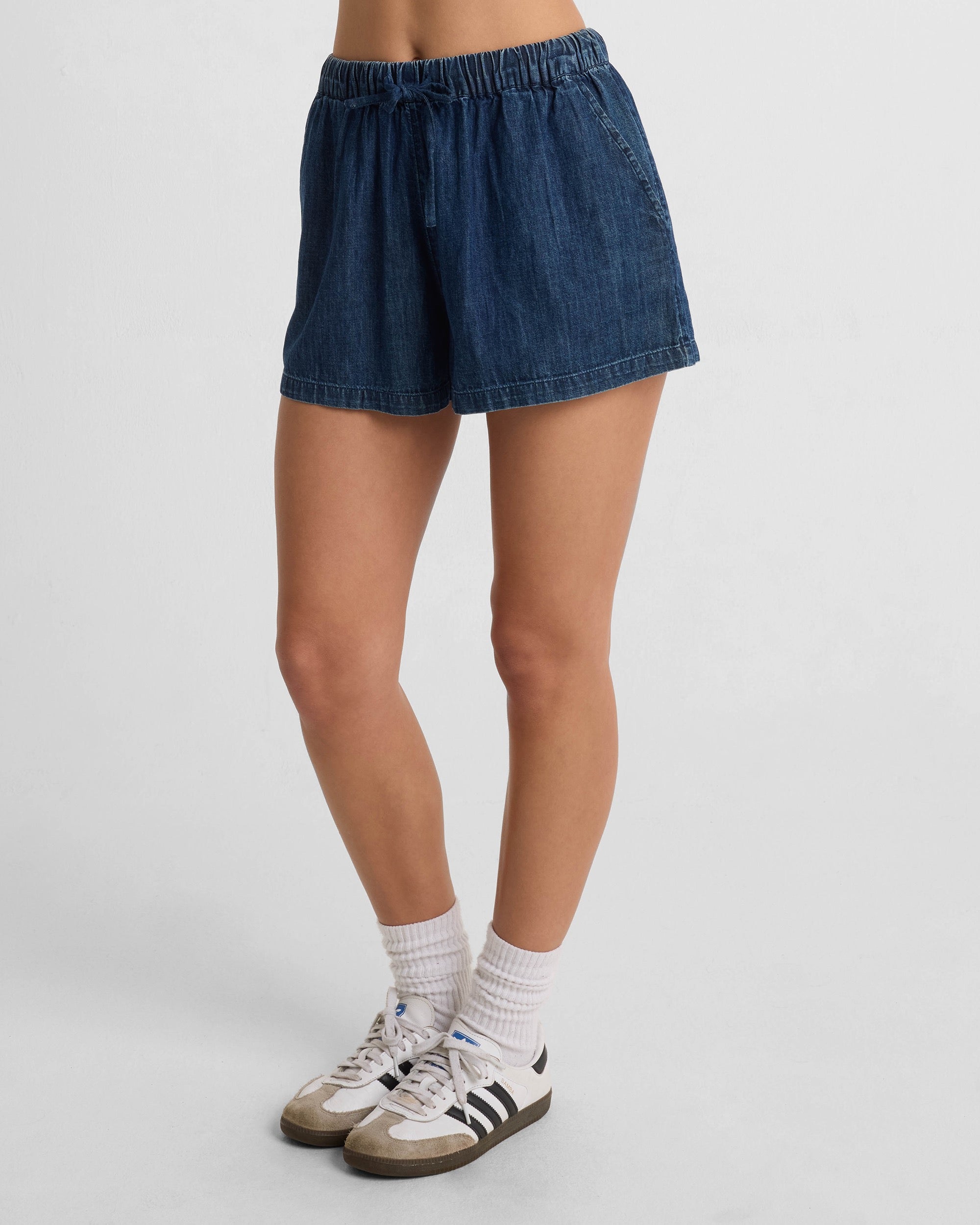 Someone stands against a plain white background wearing Volcom Women's Stone Shine Shorts in dark denim with an elastic waistband, paired with white crew socks and white athletic sneakers featuring black stripes.