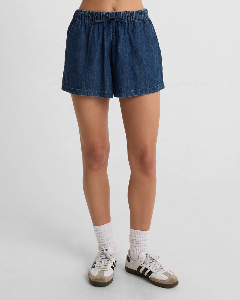 A person wears Volcom Women's Stone Shine Short with side seam pockets, white crew socks, and white-and-black sneakers against a plain white background. Only their legs and lower torso are visible.