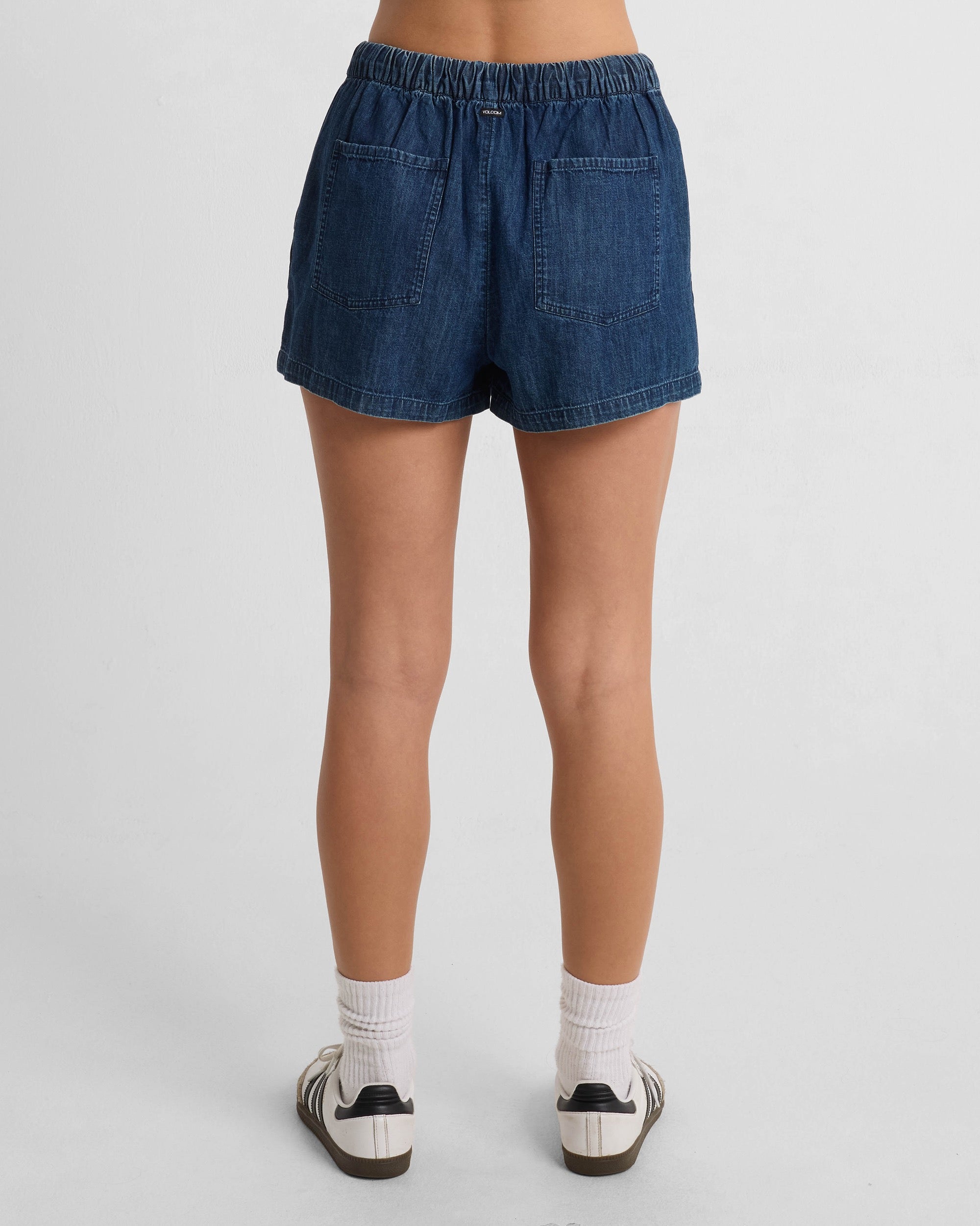 A woman faces away, wearing the Women's Stone Shine Short by Volcom in dark blue denim with side and back pockets, paired with white socks and white sneakers featuring black accents. The background is plain white.