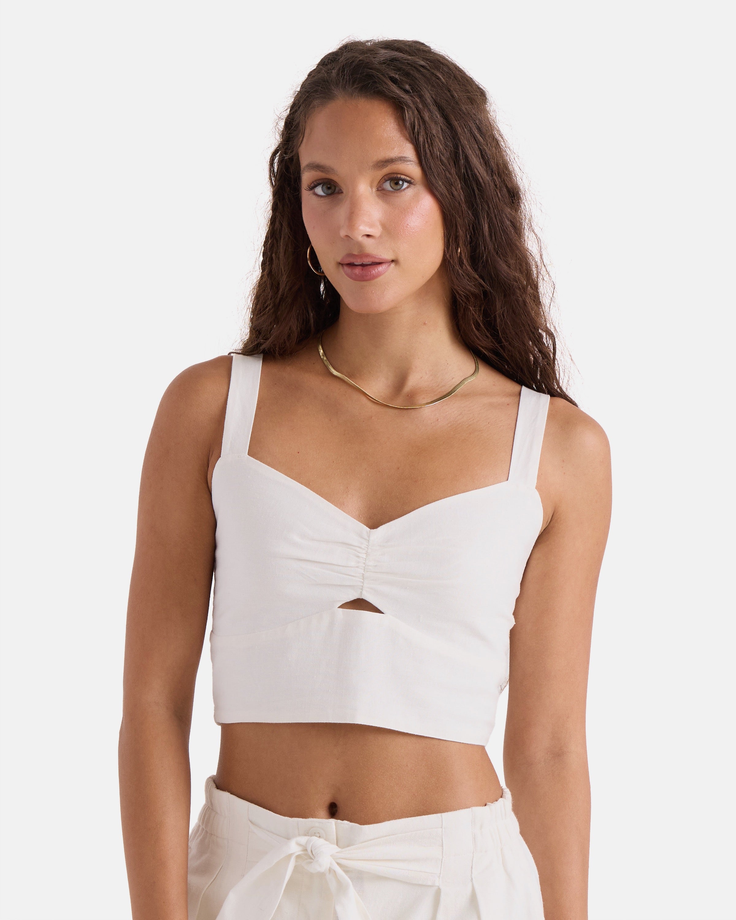 A woman with long, wavy brown hair models the Volcom Women's Bow Chicka Tank—a vintage white top with thick straps and a front cutout—paired with high-waisted white pants against a plain white background.
