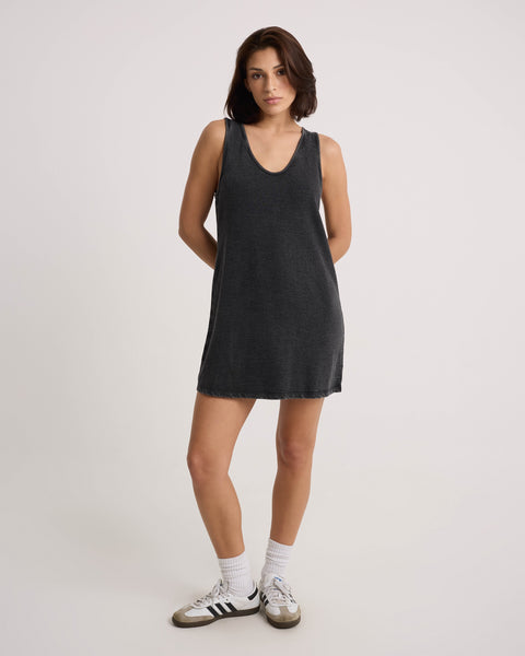 A woman with medium-length brown hair wears the Volcom Women's Glam Crop Dress, a sleeveless mini in Vintage Black, paired with white Adidas sneakers featuring black stripes, standing hands behind her back against a plain light backdrop.
