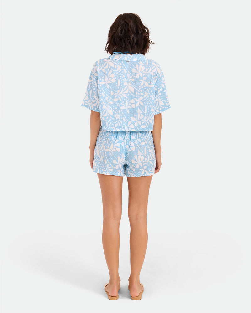 A woman with shoulder-length wavy hair faces away against a white background, wearing Volcom's Women's Funtastical Gauze Shorts—light blue and white, floral-patterned, relaxed fit with an elastic waist—paired with tan sandals.
