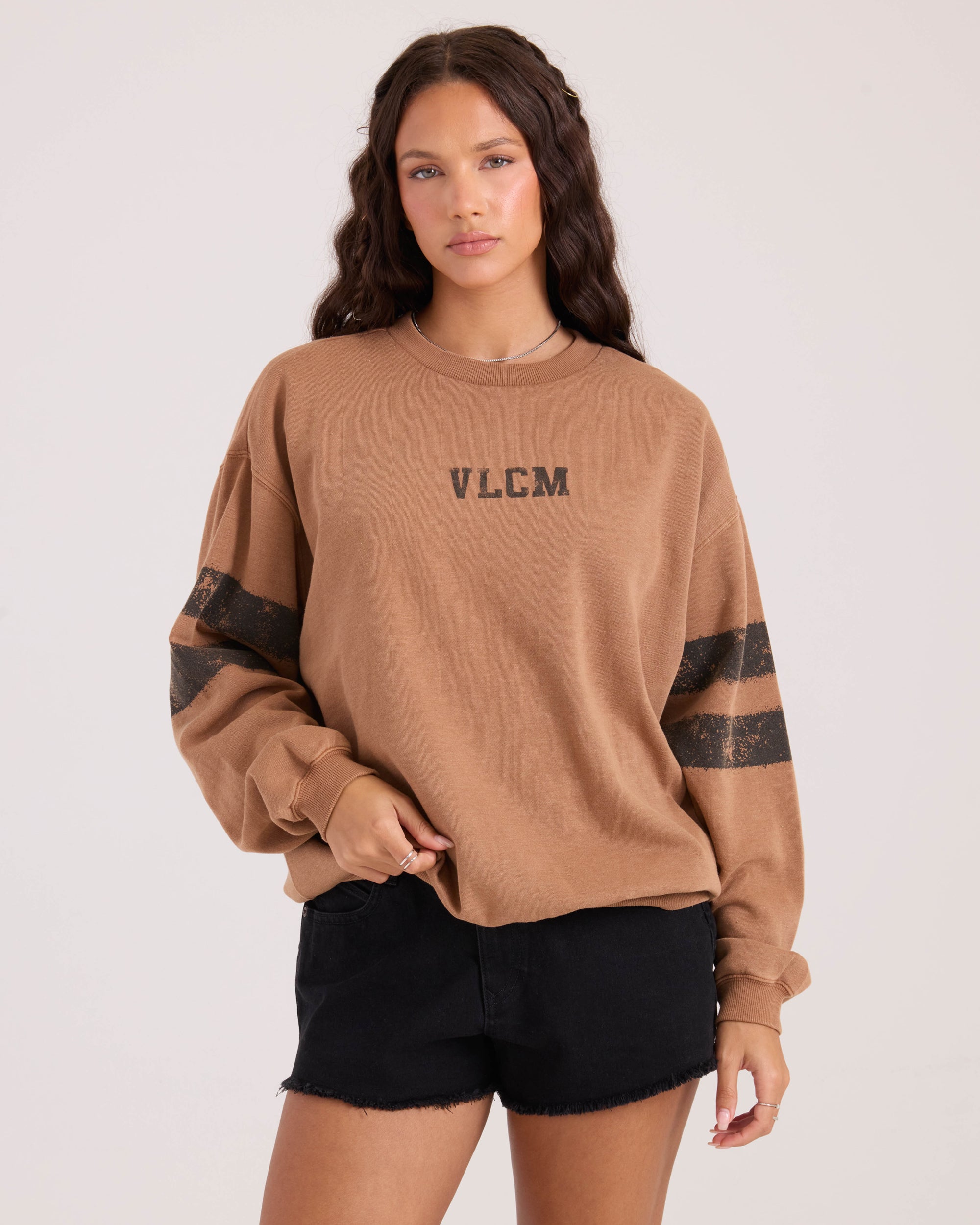 A woman with long wavy hair stands against a plain light background, wearing an oversized brown Women's Levy Crew Fleece sweatshirt by Volcom with black sleeve stripes and VLCM graphic on the front, paired with black shorts.