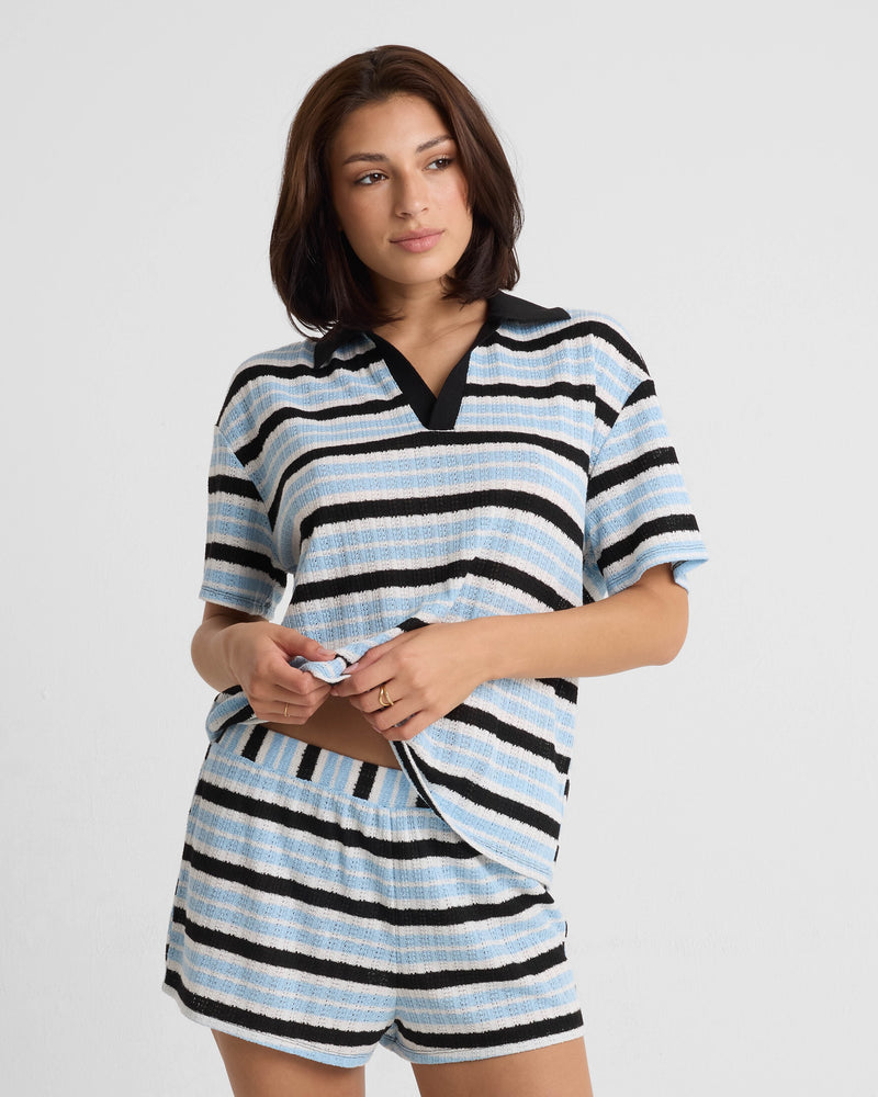 A woman with shoulder-length brown hair poses against a plain background, wearing a relaxed fit Volcom Women's Coco Ho Rugby Top in light blue, black, and white stripes. She pairs it with matching shorts and has a relaxed expression.
