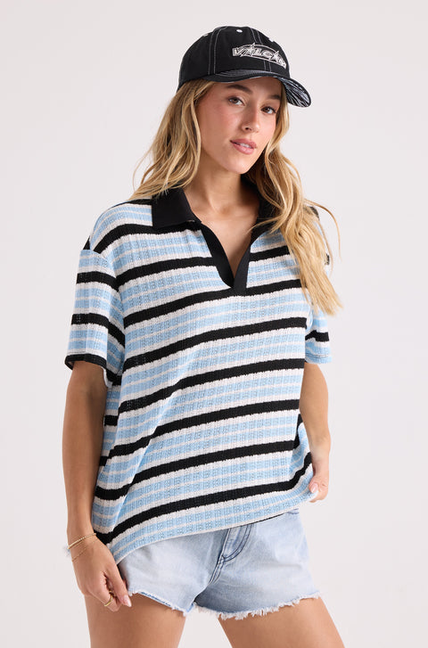 A person wears a relaxed fit Volcom Women's Coco Ho Rugby Top in blue, black, and white stripes, paired with a black baseball cap and light denim shorts against a plain background.