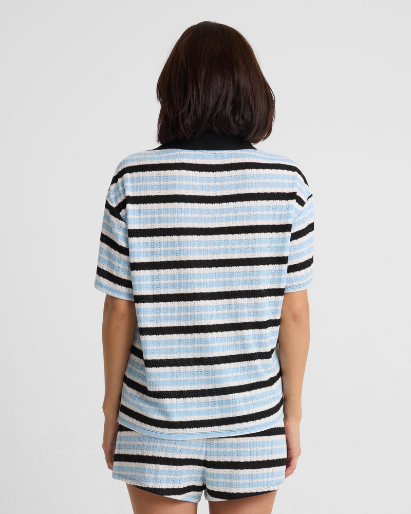 A person with shoulder-length dark hair stands with their back to the camera, wearing a relaxed fit Volcom Women's Coco Ho Rugby Top in light blue, black, and white stripes. The top features recycled polyester. Background is plain white.