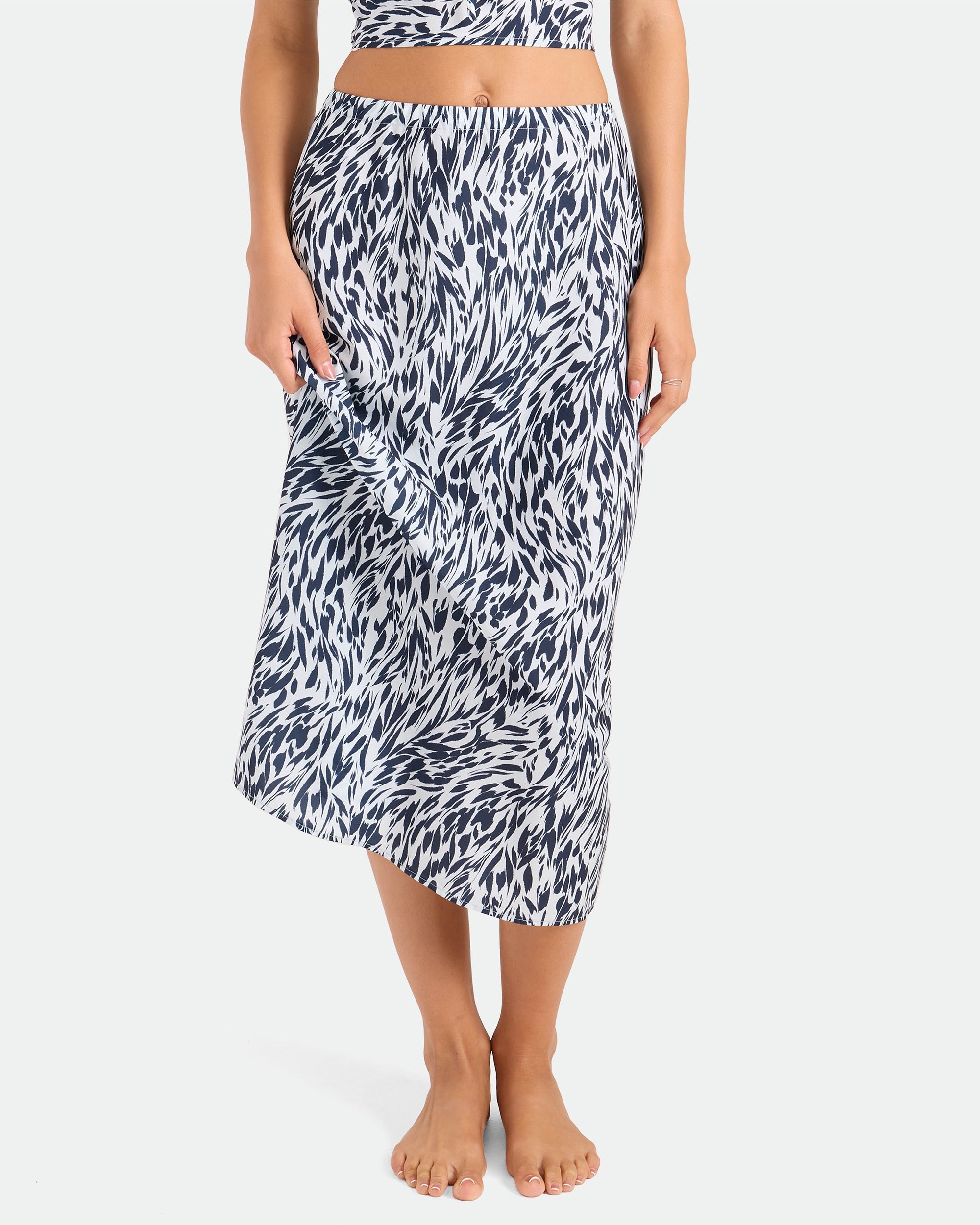A person stands barefoot against a plain white background wearing the Volcom Women's Coco Ho Sandy Parts Skirt, a high-waisted midi skirt with a woven drape and abstract brushstroke print. The person's upper body is partially visible.