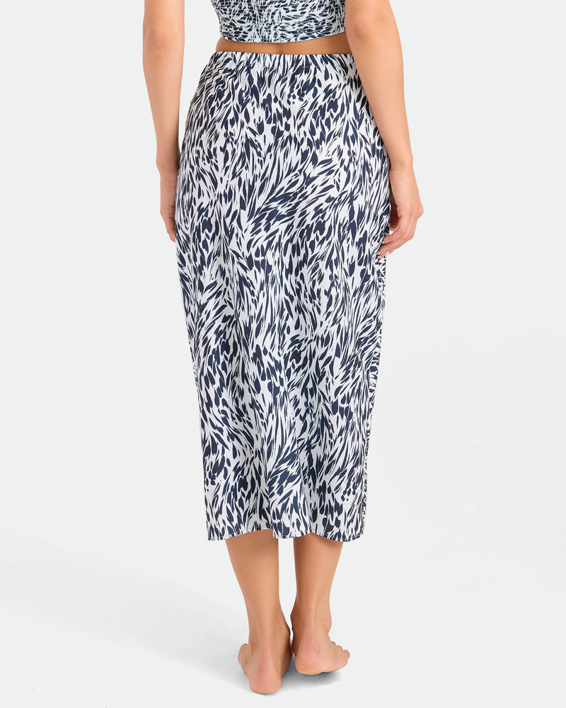 A person stands barefoot, facing away, wearing the Volcom Women's Coco Ho Sandy Parts Skirt—a high-waisted midi skirt with a black and white abstract pattern. The background is plain and light-colored.