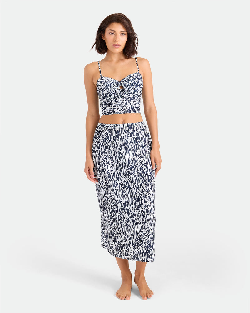 A woman stands barefoot against a white background, wearing the Volcom Women's Coco Ho Money Mama Cami—a sleeveless, cropped top—paired with a matching high-waisted, mid-length skirt featuring a blue and white abstract print.