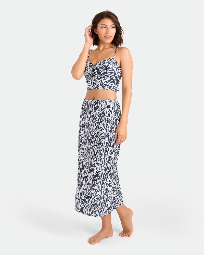 A woman with short dark hair stands barefoot in the Volcom Women's Coco Ho Money Mama Cami and matching skirt, both with a blue and white abstract leaf pattern, posing against a simple background in Coco Ho’s effortlessly stylish look.