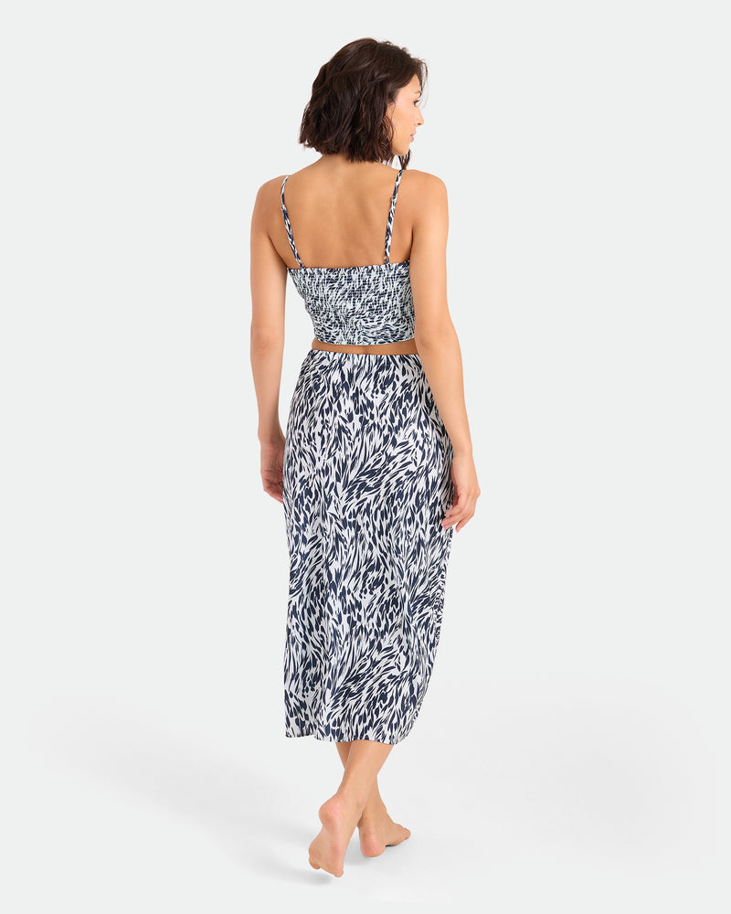 A woman stands barefoot with her back to the camera, wearing the Women's Coco Ho Money Mama Cami by Volcom—a blue and white patterned sleeveless crop top and matching high-waisted mid-calf skirt, against a plain white background.