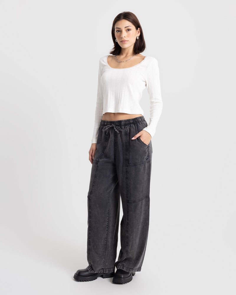 A person wears a white long-sleeved shirt and Volcom Women’s Washed Away Twill Pant, featuring vintage black, wide-leg drawstring styling with front pockets.