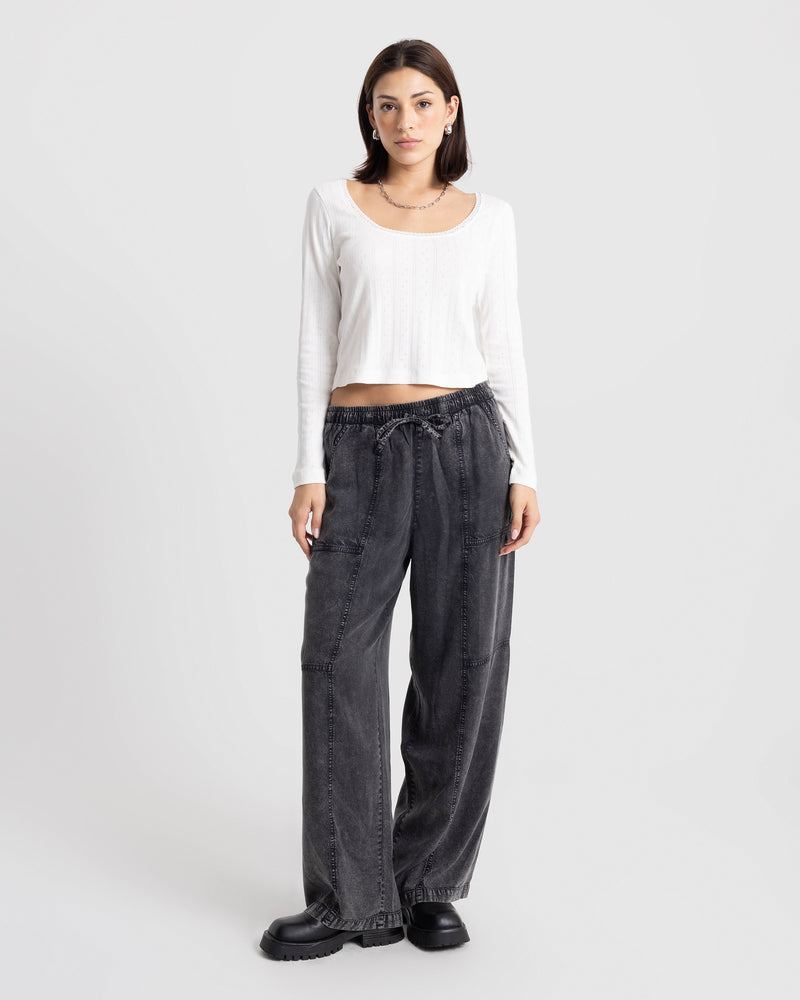 A person wears a long-sleeved white scoop-neck top and Volcom Women’s Washed Away Twill Pants in a loose-fitting, wide-leg vintage black washed style with a drawstring waist, paired with black lug-sole shoes.