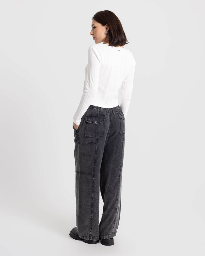 A person wearing a white long-sleeved top and Volcom Women’s Washed Away Twill Pant in charcoal grey, featuring a vintage black washed finish, a wide-leg fit, and large back pockets.