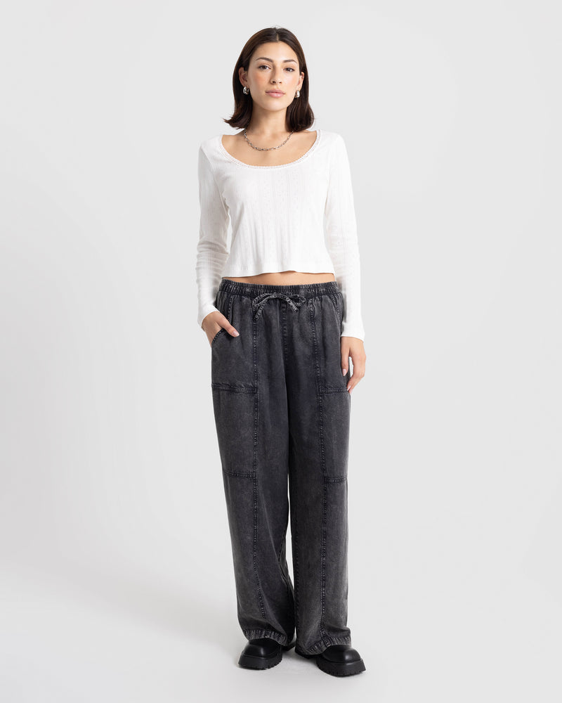 A person wears a white long-sleeve top and Volcom Women’s Washed Away Twill Pant, which are wide-leg vintage black washed pants with an elastic drawstring waist and side pockets.