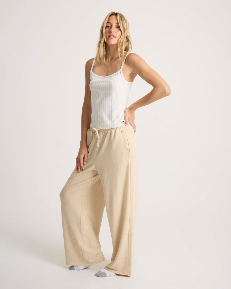 A woman stands against a plain background, wearing a white sleeveless ribbed top paired with the Volcom Women’s Lil Frenchie Pants—beige relaxed-fit lounge pants crafted from soft knit fabric with an elastic drawstring waistband. She completes her look with white socks featuring gray toes.