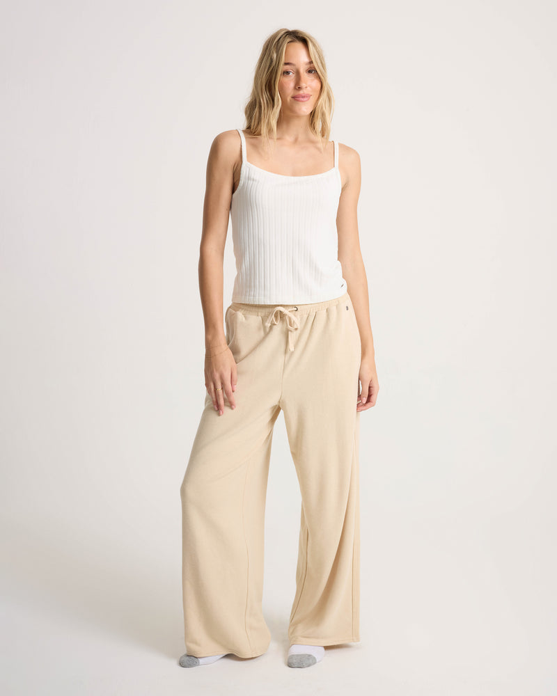 A person stands against a plain background wearing a white tank top, gray socks, and Volcom Women’s Lil Frenchie Pants in beige—featuring a relaxed fit and elastic waistband—smiling slightly with one hand resting at their side.