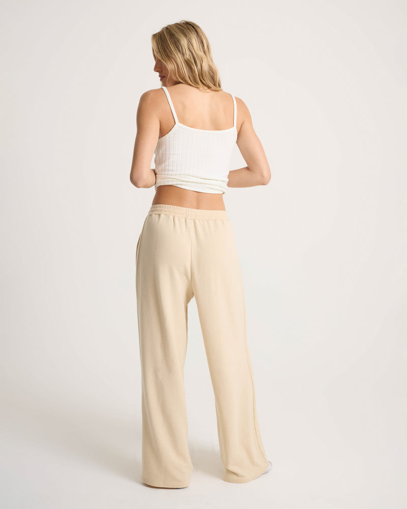 A person with long blonde hair stands facing away, wearing a white ribbed tank top and Volcom Women’s Lil Frenchie Pants in a loose beige relaxed fit with an elastic waistband against a plain light background.