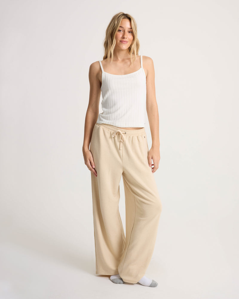 A person stands on a plain background wearing Volcom’s Women’s Lil Frenchie Pants in beige, featuring a relaxed fit and elastic waistband, paired with a white tank top. They have long, wavy hair and are styled with gray and white socks for a comfortable casual look, enhanced by the pants’ soft knit fabric.