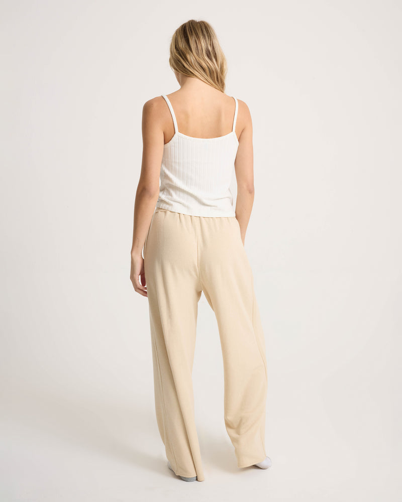 A person with long blonde hair, wearing a white tank top and Volcom’s Women’s Lil Frenchie Pants in a relaxed beige fit with an elastic waistband, stands facing away against a plain white background.