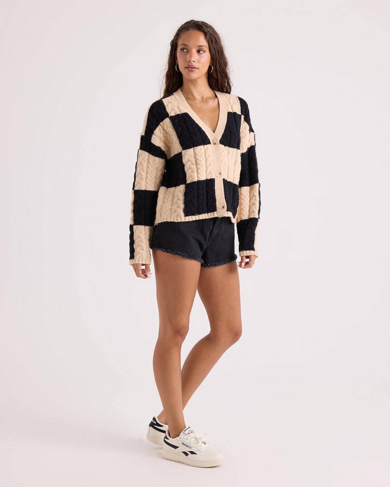 A woman with long wavy hair models the Volcom Women's Check You Sweater—a beige and black checkerboard patterned V-neck cardigan with a comfy knit and button front—paired with black denim shorts and white sneakers, standing against a plain white background.