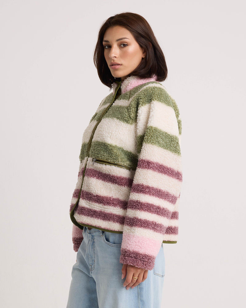 A woman with a bob haircut models the Volcom Women’s Sherpa Stripe Jacket, showcasing plush sherpa fabric in green, white, and purple stripes. She pairs the boxy-fit jacket with light blue jeans and stands sideways against a plain white background, highlighting its modern silhouette.
