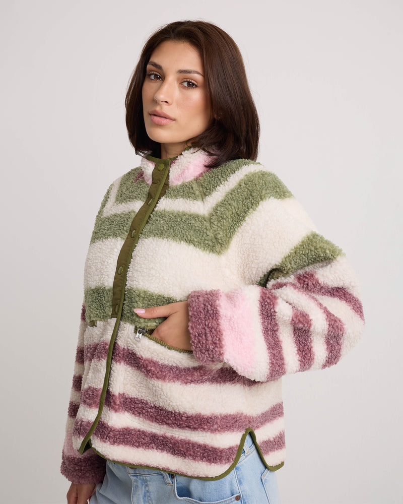 A woman with shoulder-length brown hair models the Volcom Women’s Sherpa Stripe Jacket, showcasing its green, white, and pink striped design. The plush sherpa material and boxy fit give this jacket a cozy, relaxed appearance as she stands with one hand in her pocket against a plain light background.