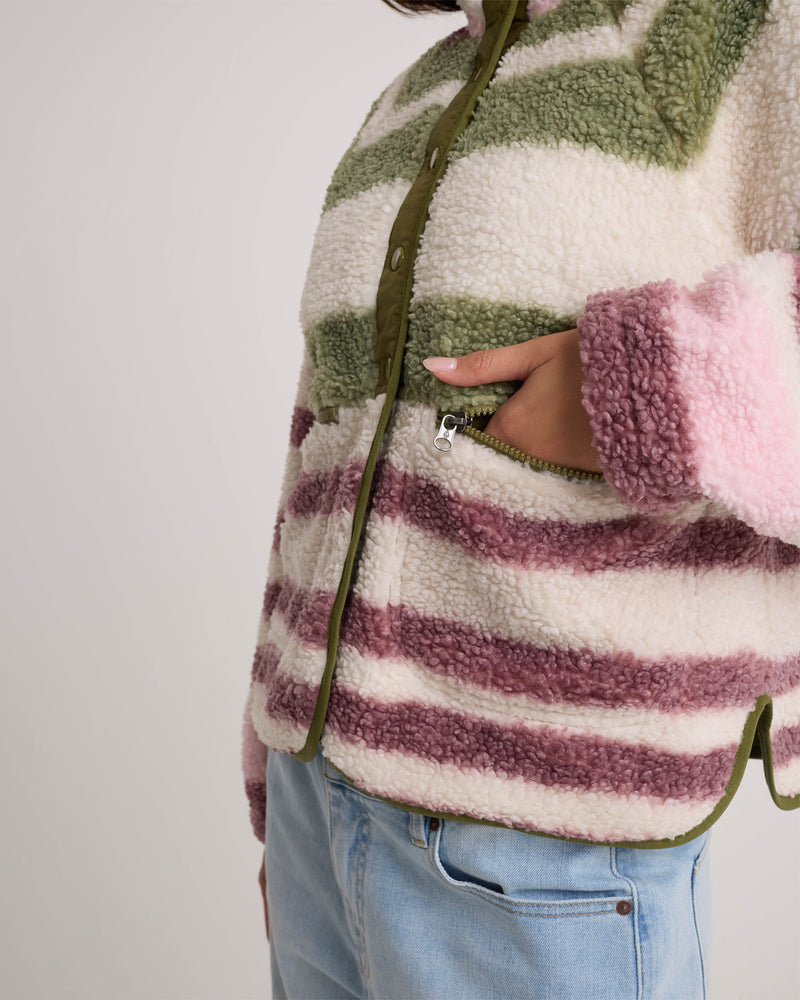 A person wears the Volcom Women’s Sherpa Stripe Jacket, featuring plush sherpa material in green, pink, and purple stripes and a cozy, boxy fit. One hand rests in a zippered pocket. The lower part of their face is not visible. They are also wearing light blue jeans.