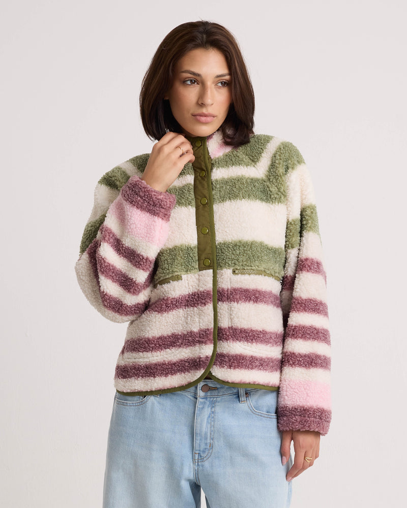 A woman wears the Volcom Women’s Sherpa Stripe Jacket, featuring shades of pink, green, and white with a boxy fit and plush sherpa texture, paired with blue jeans and standing against a plain white background.