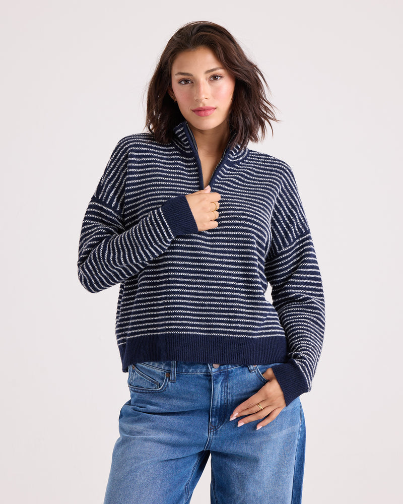 A woman with shoulder-length dark hair wears the Volcom Women's Sun Of Sand Sweater—a navy and white striped quarter-zip design—paired with blue jeans, standing against a plain light background with one hand touching her collar.