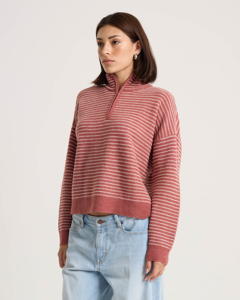 A woman wears the Volcom Women’s Sun Of Sand Sweater—a pink and white striped pullover with a half-zip collar and boxy fit—paired with light blue jeans, standing against a plain, light background.