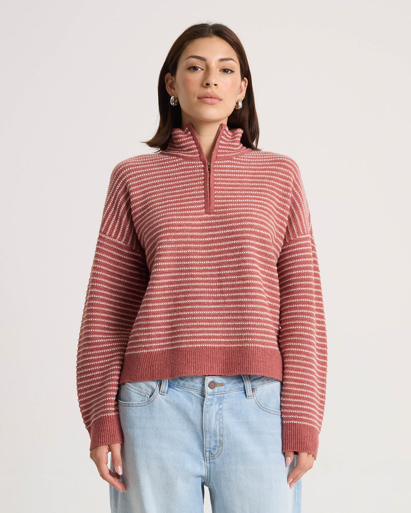 A woman stands facing forward in front of a plain light background, wearing the Volcom Women’s Sun Of Sand Sweater—a pink and white striped pullover with a half-zip collar and boxy fit—paired with light blue jeans.