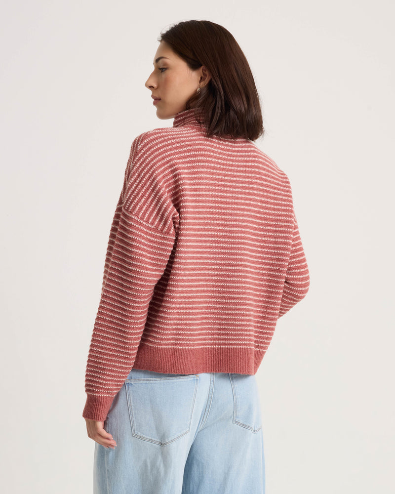 A woman with short brown hair is seen from behind, standing against a plain background and wearing the Volcom Women’s Sun Of Sand Sweater—a pink and white striped pullover turtleneck with a boxy fit—paired with light blue jeans.
