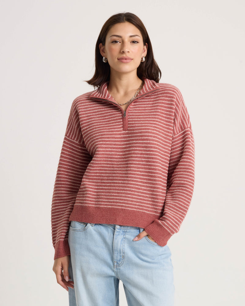 A woman stands against a plain light background with one hand in her pocket, wearing the Volcom Women’s Sun Of Sand Sweater—a pink and white striped pullover with a boxy fit—paired with light blue jeans.