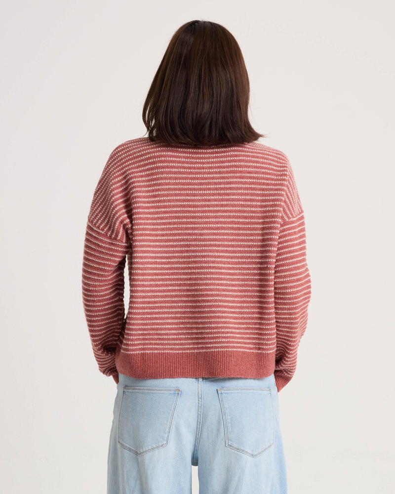 A person with shoulder-length brown hair is standing with their back to the camera, wearing the Volcom Women’s Sun Of Sand Sweater—a pink and white striped pullover with a boxy fit—paired with light blue jeans against a plain light background.