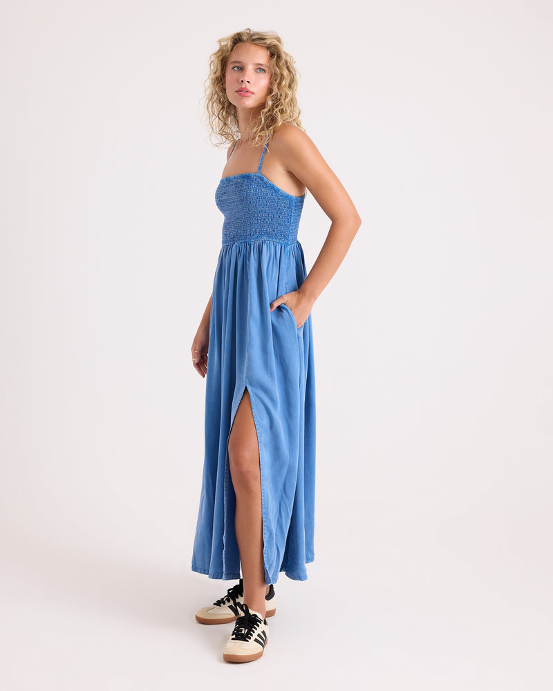 A woman stands against a plain white background wearing the Volcom Women’s Santa Monica Beach Maxi—a blue, sleeveless viscose maxi dress featuring an ankle-length hem, side slit, and convenient pockets. She pairs the look with black and white sneakers.