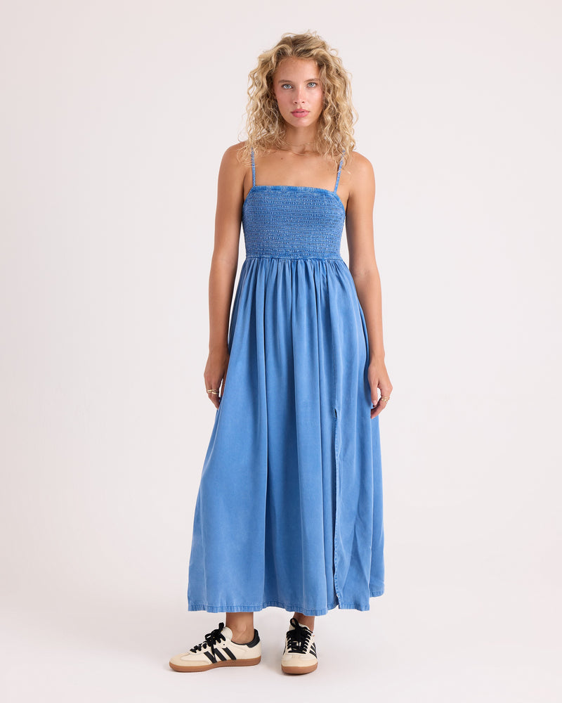 A woman with curly blonde hair wears the Volcom Women’s Santa Monica Beach Maxi dress in blue, styled with white sneakers featuring black stripes, as she stands against a plain light background.