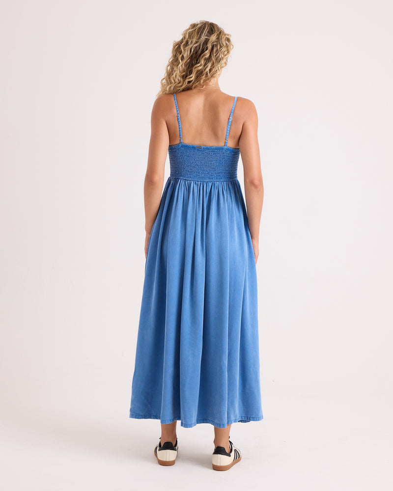 A woman with curly blond hair stands facing away, wearing the Volcom Women’s Santa Monica Beach Maxi and black-and-white sandals, against a plain white background.